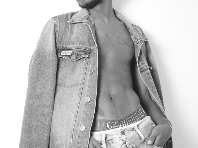 AlexJaccob's shirtless torso with a denim jacket, showing a tattoo on his chest, and wearing unbuttoned jeans with "Guess" underwear, black and white image.