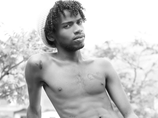 Black-and-white photo of AlexJaccob, shirtless with curly hair, tattoos on chest, and a serious expression. He's wearing a white hat, and his eyes are focused.