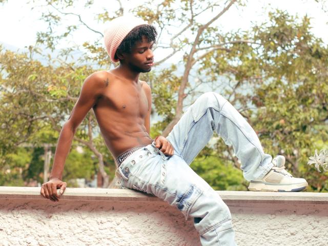 shirtless AlexJaccob with a slim, toned, dark brown body, wearing a pink beanie, light blue jeans, and white sneakers, sitting on a white wall, looking down.