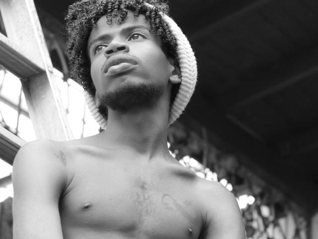Black and white photo of AlexJaccob, shirtless with curly hair, beanie, and small nipple, looking up with a pouty face, tattoos on chest, under a roof.