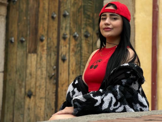 JulietaDupont, with black hair, red cap, red tank top, black and white fuzzy jacket, and necklace, sits smiling, showing her tits, against a wooden door.