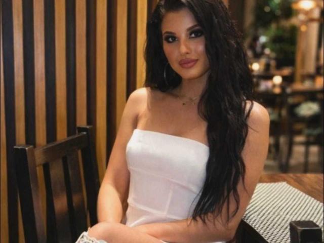 KatherinWow with long black hair, wearing a white strapless top, sitting with arms crossed, showing her tits, in a dimly lit room with wooden vertical panels.