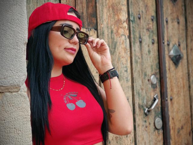 JulietaDupont with black hair, red cap, red sleeveless top, brown sunglasses, black wristwatch, and red lipstick, adjusting sunglasses, showing tattoos on her right arm.