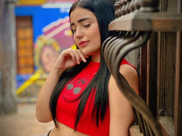 JulietaDupont with long black hair, red crop top, and medium tits, leaning against wooden structure, hand near mouth, soft smile, slightly blurred background, colorful wall art behind.