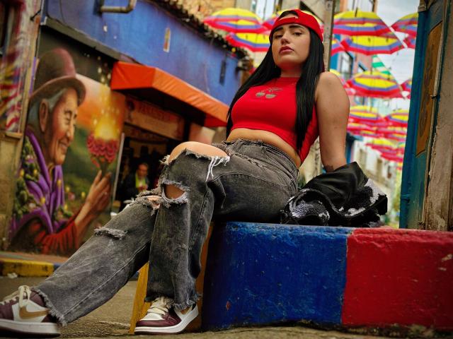 JulietaDupont with black hair, red headband, red crop top, and ripped jeans, sitting on blue and red block, looking annoyed, with black jacket on her lap.