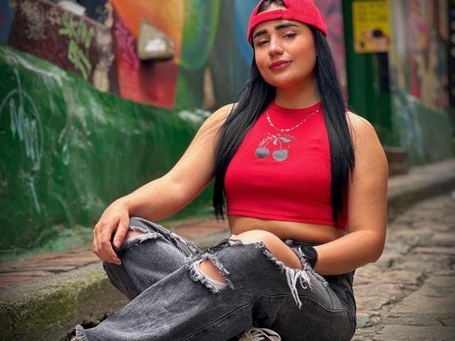 JulietaDupont, with long black hair, wearing a red cap and crop top, showing her tits in a red top, and ripped jeans, sitting on a cobblestone street.