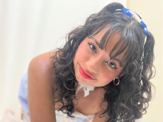 AsleyLoren has curly black hair, blue headband, red lipstick, hoop earrings, and is looking up with a smile, wearing a white choker and blue top, showing her tits.