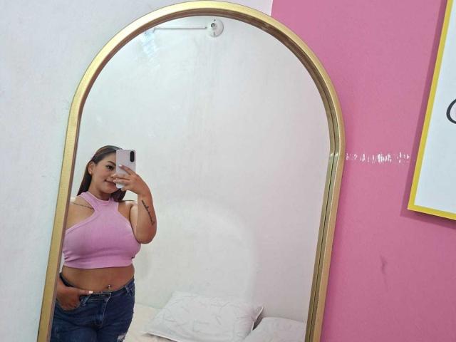 KinkyWildQueen with medium tits, wearing a pink crop top and blue jeans, taking a mirror selfie, smiling, hands in pockets, in a pink and white room.
