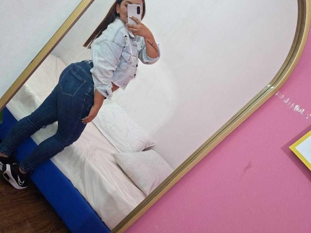 KinkyWildQueen macht ein Selfie vor einem Spiegel, trägt eine Jeansjacke, enge Jeans und schwarze Schuhe und präsentiert dabei ihr kurviges Hinterteil und ihre kleinen Brüste. Sie hält ein Handy..