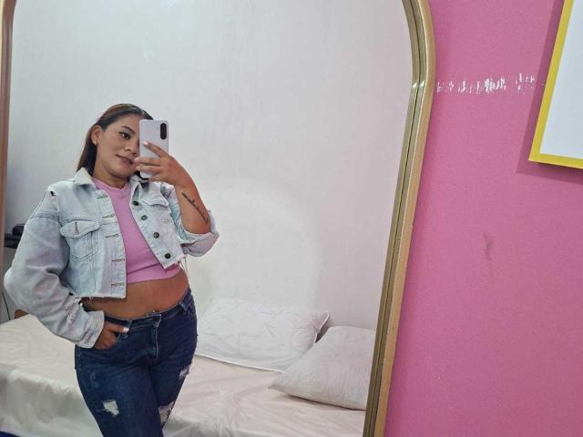 KinkyWildQueen, mit einem süßen Lächeln, steht vor einem Spiegel und trägt ein pinkes Crop-Top, zerrissene Jeans und eine Jeansjacke, wobei ihr schlanker, kurviger Hintern zu sehen ist..