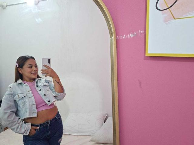 KinkyWildQueen with pink hair, wearing a denim jacket and pink top, shows her flat stomach and hand in her jeans, taking a mirror selfie in a pink room.