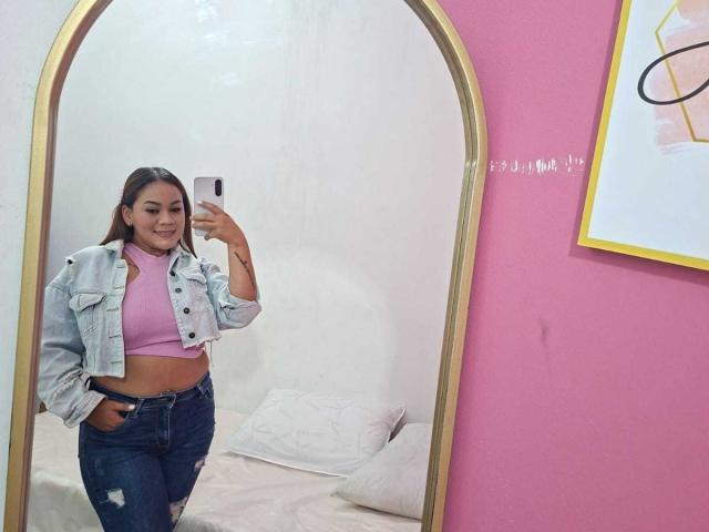 KinkyWildQueen, con la pelle abbronzata, i capelli castani, indossa una canotta rosa, una giacca di jeans e jeans strappati, è in piedi di fronte a uno specchio, tiene in mano un telefono e sorride..