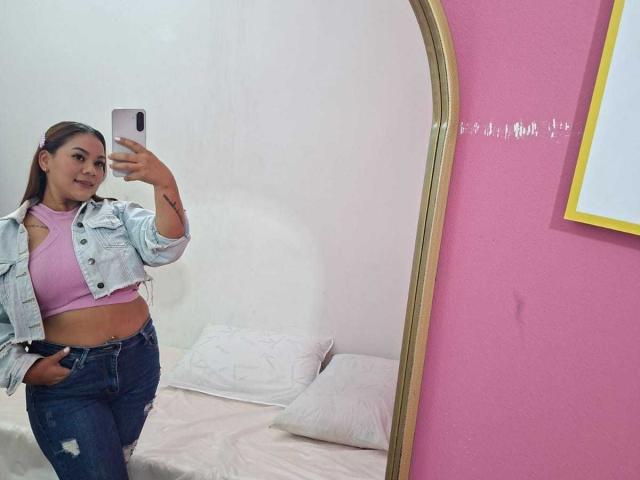 KinkyWildQueen con cabello castaño, una blusa rosa, una chaqueta vaquera y jeans, tomando una selfie frente al espejo, mostrando sus senos, con una sonrisa, en una habitación con paredes rosas..