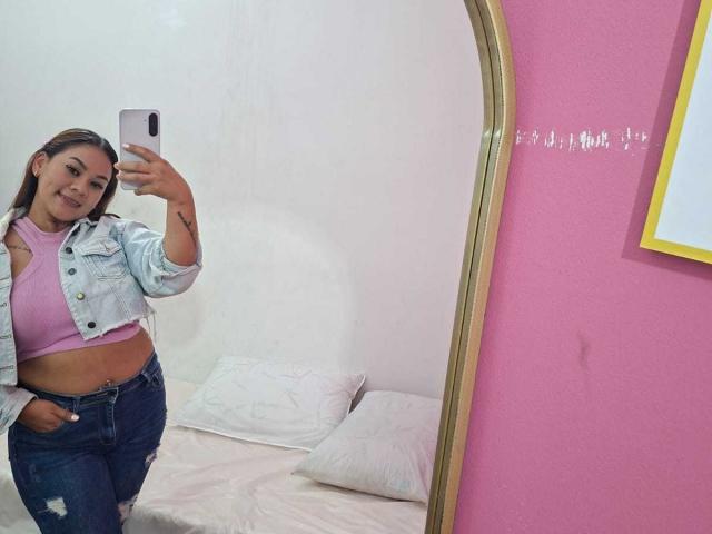 KinkyWildQueen con i capelli castano chiaro, indossa una canotta rosa, una giacca di jeans e jeans strappati, sta scattando un selfie allo specchio, mostrando il sedere e un tatuaggio sul braccio destro..