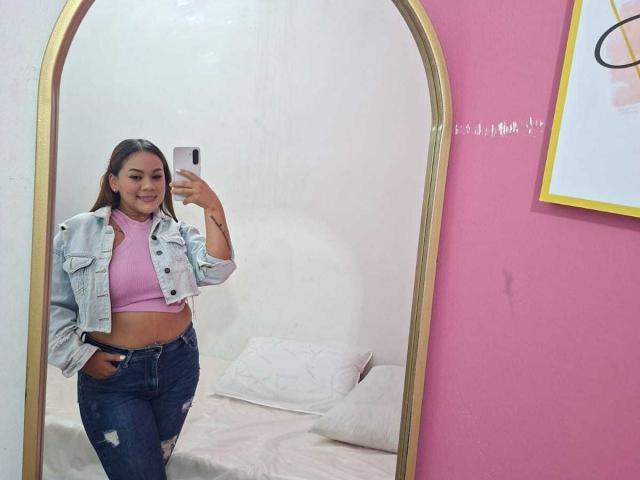 KinkyWildQueen with a cute smile, wearing a pink turtleneck, denim jacket, and ripped jeans, standing in front of a mirror, holding a phone, showing her midriff and ass.