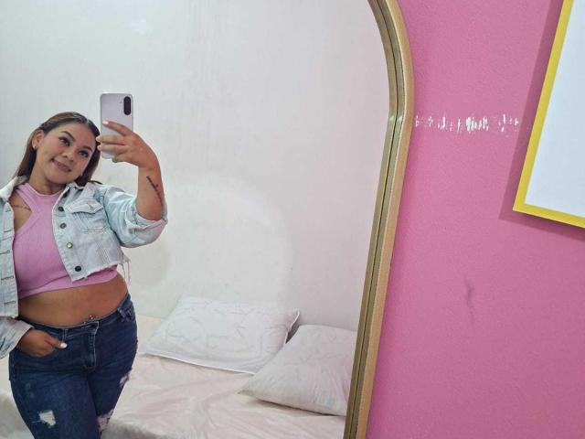 KinkyWildQueen com cabelo castanho, usando um sutiã rosa, jaqueta jeans e calças jeans rasgadas, tirando uma selfie no espelho, mostrando seus seios pequenos e a tatuagem no umbigo..