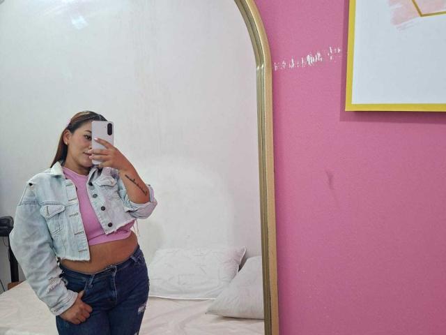 KinkyWildQueen draagt een roze topje en gescheurde jeans, met een denimjasje, en maakt een spiegelzelfportret met een glimlach, waarbij haar kleine achterwerk en tatoeage op haar rechterarm zichtbaar zijn..