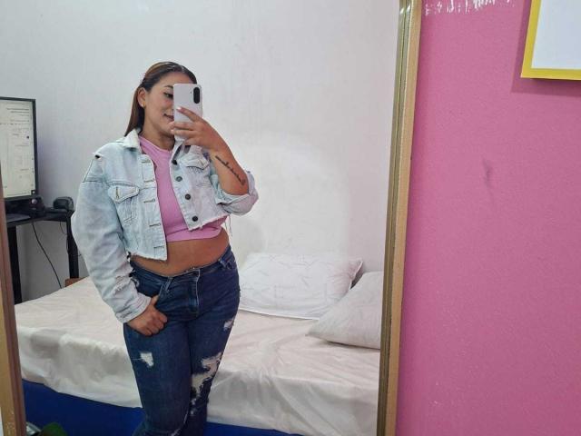 KinkyWildQueen, avec une peau bronzée, des cheveux bruns, portant un haut rose, une veste en jean et un jean déchiré, prenant un selfie devant un miroir, montrant son ventre plat et son tatouage sur la main..
