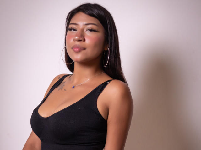 BonnieBond, with dark hair, wearing a black tank top, showing her tits, hoop earrings, and a necklace, pouting slightly, standing against a plain background.