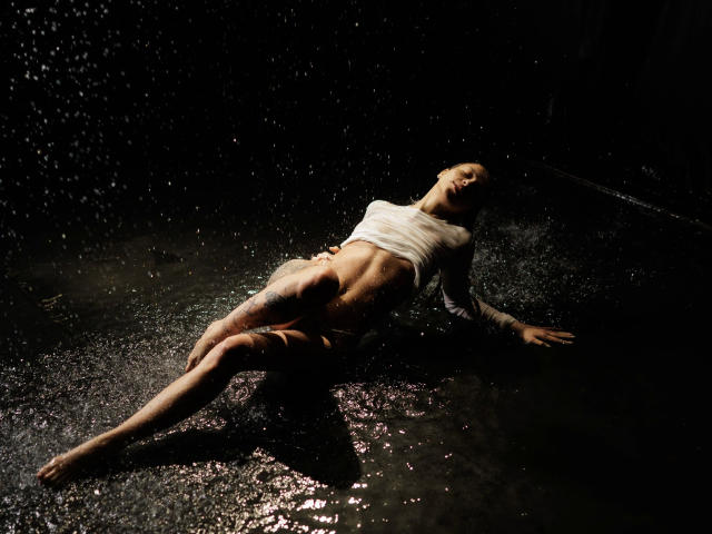 Helena-hot, wet and sexy, laying on dark, wet floor, wearing white shirt, exposing her ass, back, and tattooed legs, with water droplets, head tilted, eyes closed, arms spread.