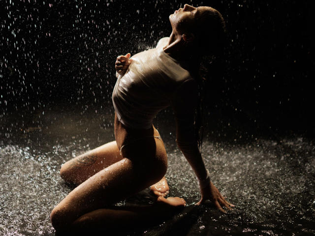 Helena-hot kneels on wet ground, back arched, white shirt clinging to her tits, water droplets everywhere, head tilted up, dark background, intense lighting, wet skin, sexy pose.