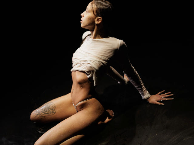 Helena-hot with wet shirt, exposing tits, sitting on knees in water, white thong, thigh tattoo, dark background, wet skin, raised arms, side profile, looking up, sexy pose.