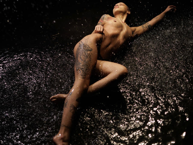 Nude Helena-hot with tattoos, wet skin, and small tits, lying in dark water, legs crossed, arm raised, water splashing, head tilted back, looking relaxed and sexy.