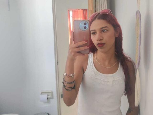 ValerieWoods with red hair, white tank top, and red lipstick, holding phone, wearing bracelets, necklace, and pink sunglasses, in mirror selfie. Boobs visible. Red lipstick.