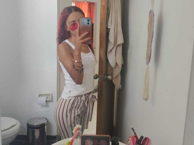 ValerieWoods with red hair, wearing a white tank top and striped pajama pants, holding a phone, showing her ass in a mirror, in a bathroom.