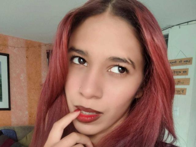 ValerieWoods with red hair, brown eyes, nose ring, and red lipstick, biting her finger, showing her glossy lips. She has a cute face and a slight smirk.