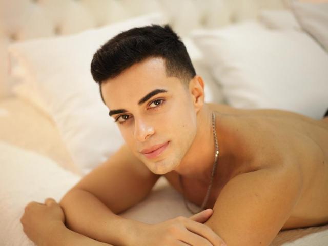 HunterParker, shirtless, with short black hair, lying on a bed, arms crossed, wearing a necklace, brown eyes, slight smile, tan skin, looking at camera.
