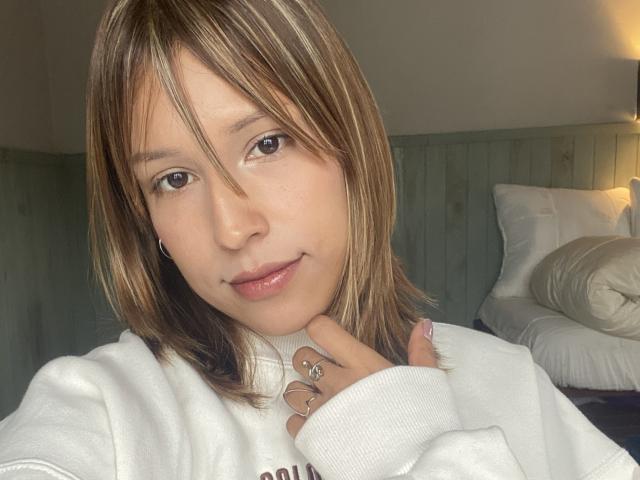 KiaraHernandezX with shoulder-length brown hair, light skin, subtle makeup, wearing a white sweatshirt, holding her neck, smiling, ring on finger, soft lighting, bedroom setting.