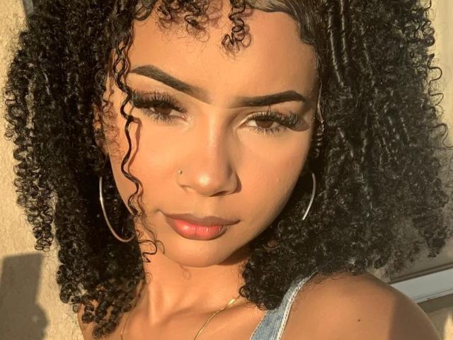 HotSweetHard with curly black hair, tan skin, thick eyebrows, long eyelashes, hoop earrings, nose piercing, and pink lips, looking down with a subtle smile in sunlight.