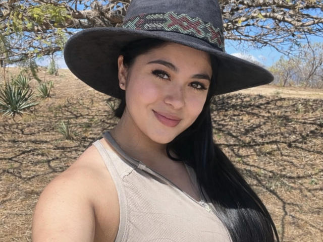 CataleyaHottyX with long black hair, tan skin, and small tits, wearing a gray hat with a patterned band, and a beige tank top, smiling outdoors.