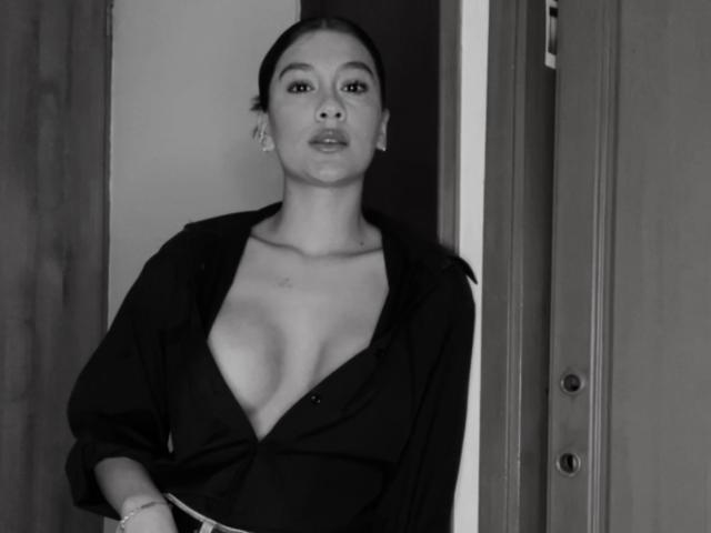 ThaliaAres has dark hair, wearing a black shirt open to reveal her tits, standing with a neutral expression in a grayscale photo. She's looking directly at the camera.