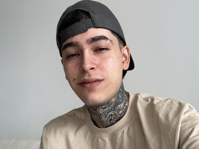 Samxx, con una gorra gris, una camisa marrón claro y un tatuaje negro detallado en el cuello, tiene el pelo corto y oscuro y una ligera sonrisa. Su piel es clara y con pecas..