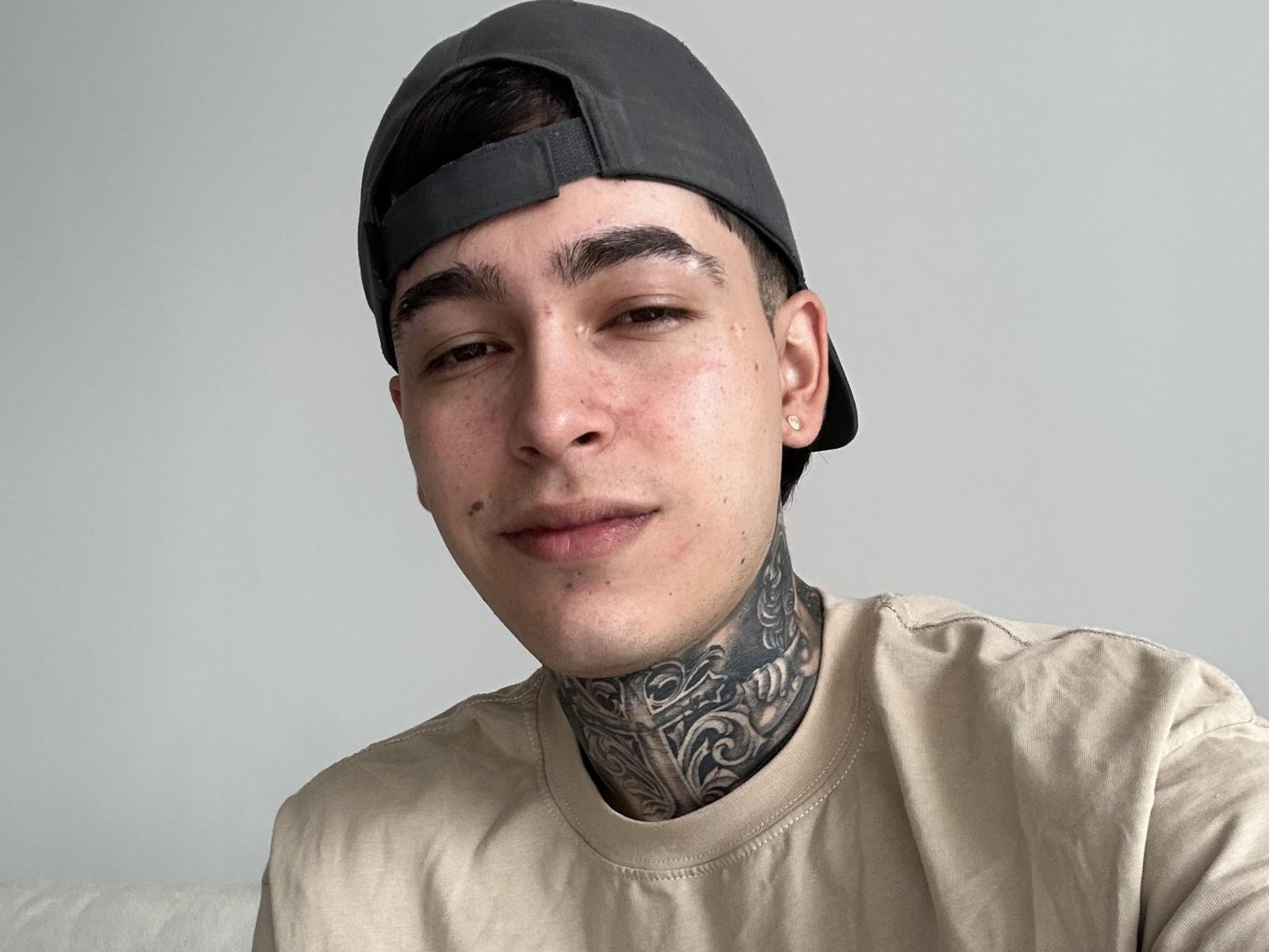 Samxx, mit einer grauen Mütze, einem hellbraunen Hemd und einem detaillierten schwarzen Tattoo im Nacken, hat kurze, dunkle Haare und ein leichtes Lächeln. Seine Haut ist hell und mit Sommersprossen übersät..