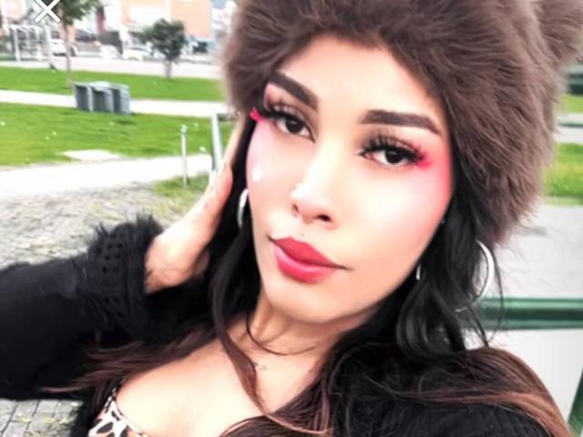 LexieBloom with pink eyeliner, red lipstick, brown fur hat, black fur-lined jacket, and hoop earrings, showing off her cute face and medium tits. She's holding her head.