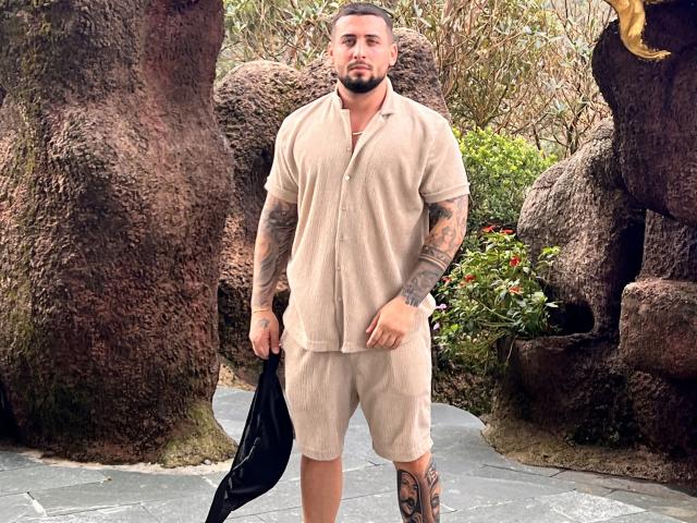 GreenDylan stands with a serious look, wearing a beige button-up shirt and shorts, holding a black bag. He has tattoos on his arms and a leg.