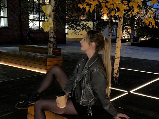 RuthaLoil with long blonde ponytail, sitting on wooden bench, wearing gray leather jacket, black shorts, black stockings, black shoes, holding coffee cup, under yellow-leaved tree at night.