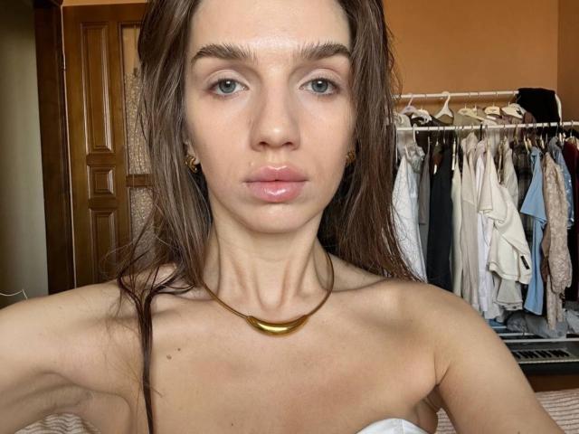 Portrait of PortiaKomer with wet brown hair, green eyes, shiny pink lips, small gold necklace, and bare shoulders, showing her tits. She's in a bedroom with clothes.