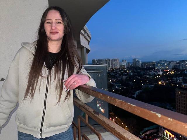 Omella stands on a balcony at dusk, wearing a beige zip-up jacket and blue jeans, with long brown hair and a subtle smile, holding a rusty railing.