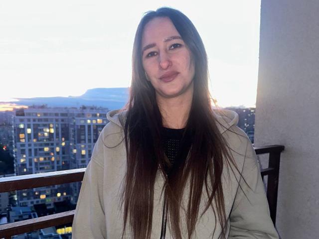 Omella with long brown hair, light green jacket, and subtle smile, sitting on balcony at dusk, city lights in background, her ass barely visible under jacket.