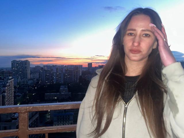 Omella with long brown hair, wearing a white zip-up jacket, saluting with her right hand, sunset in the background, city skyline visible, blue and orange sky, urban setting.