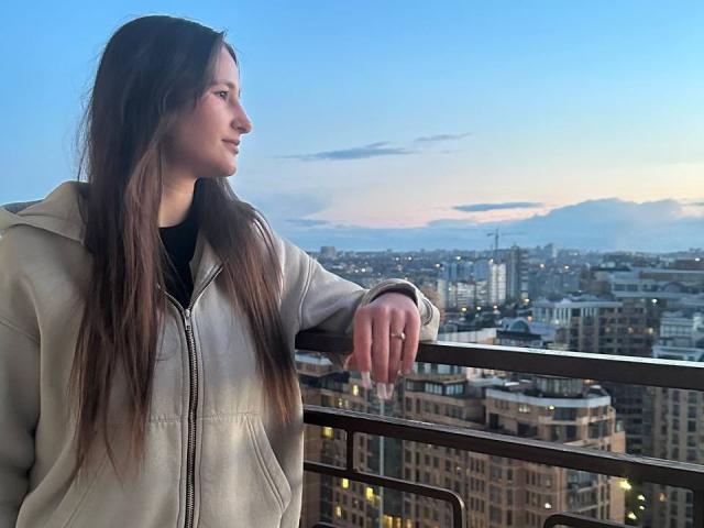 Omella with long brown hair, wearing a beige jacket, leans on a balcony railing, looking to the side, city skyline in the background at dusk. Her hand rests on the rail.