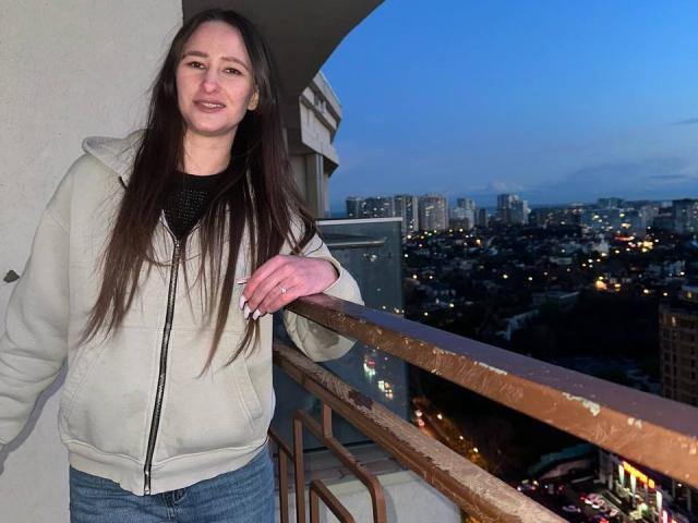 Omella stands on a balcony, wearing a beige zip-up hoodie and blue jeans, with long brown hair, and a slight smile, during dusk. Her hand rests on a rusted railing.
