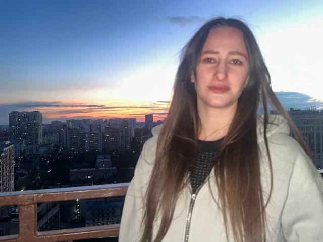 Omella with long brown hair, wearing a gray hoodie, standing on a balcony at sunset, city skyline in the background, face slightly puffy, hair blowing in the wind.