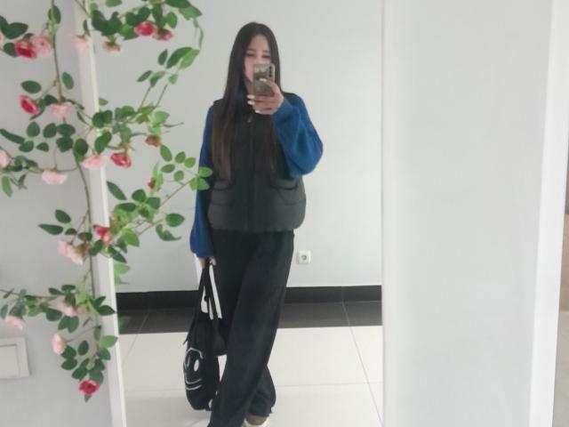 Omella with long black hair, wearing a blue jacket, black vest, and black pants, holding a black bag, taking a mirror selfie. Pink rose vine in the mirror.