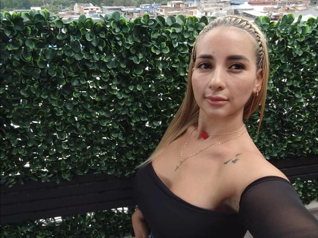 Lanna Brown met blonde vlechten, een zwarte top met een open schouder, kleine borsten, een gouden ketting, een rode hanger, een tatoeage op de rechterkant van de borst, een groene, bladerige achtergrond, een lichte glimlach, lichte huid, een selfie..