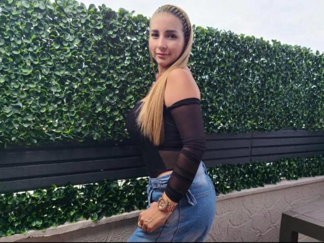 Lanna Brown, con trecce bionde, una maglia nera trasparente, jeans blu e un orologio dorato, è in piedi accanto a una panchina nera, sorridendo alla fotocamera, mostrando i glutei e i seni piccoli..